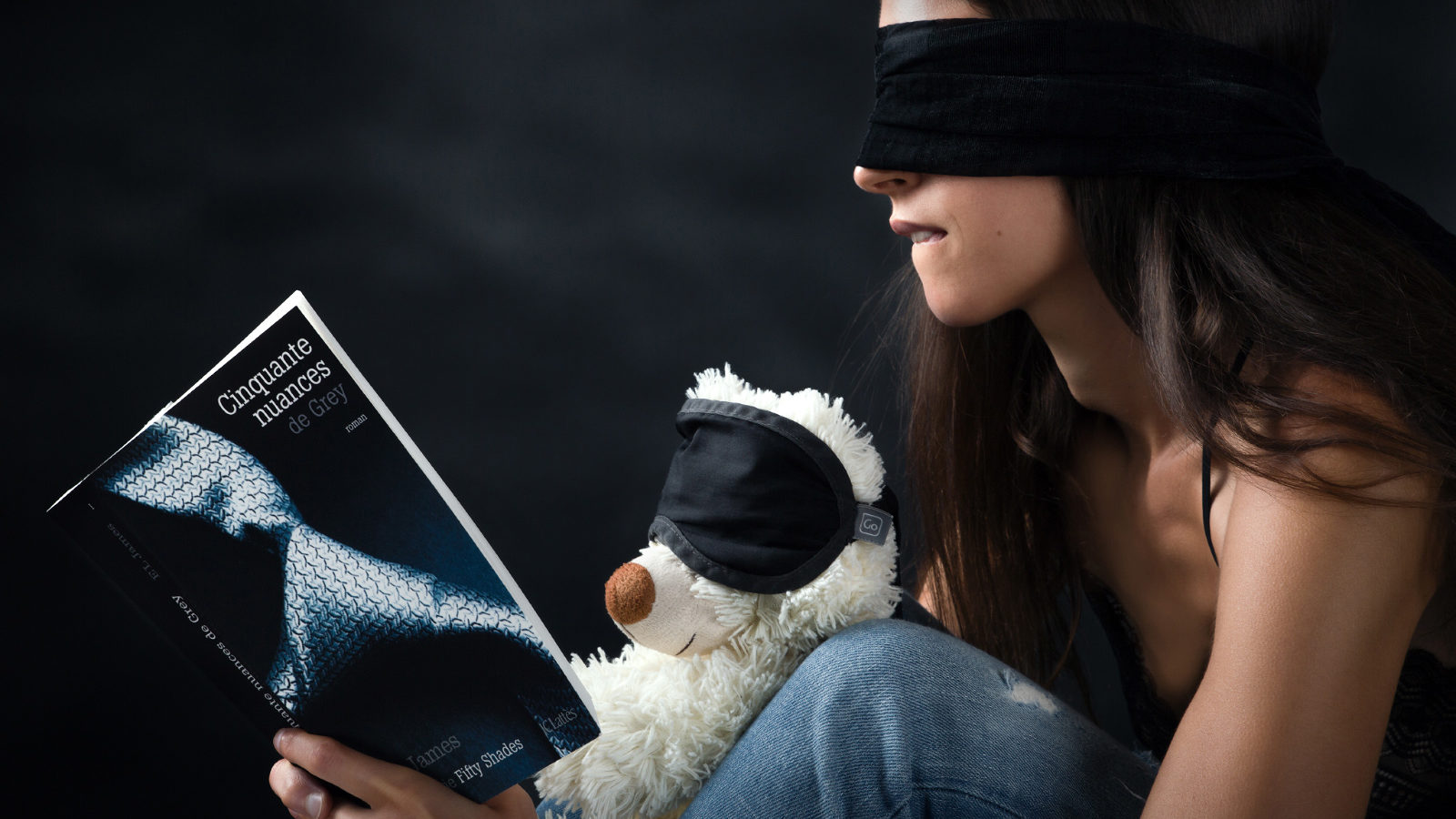 Un ours en peluche et un modèle photo, les yeux bandés, semblent apprécier le livre 50 Nuances de grey.