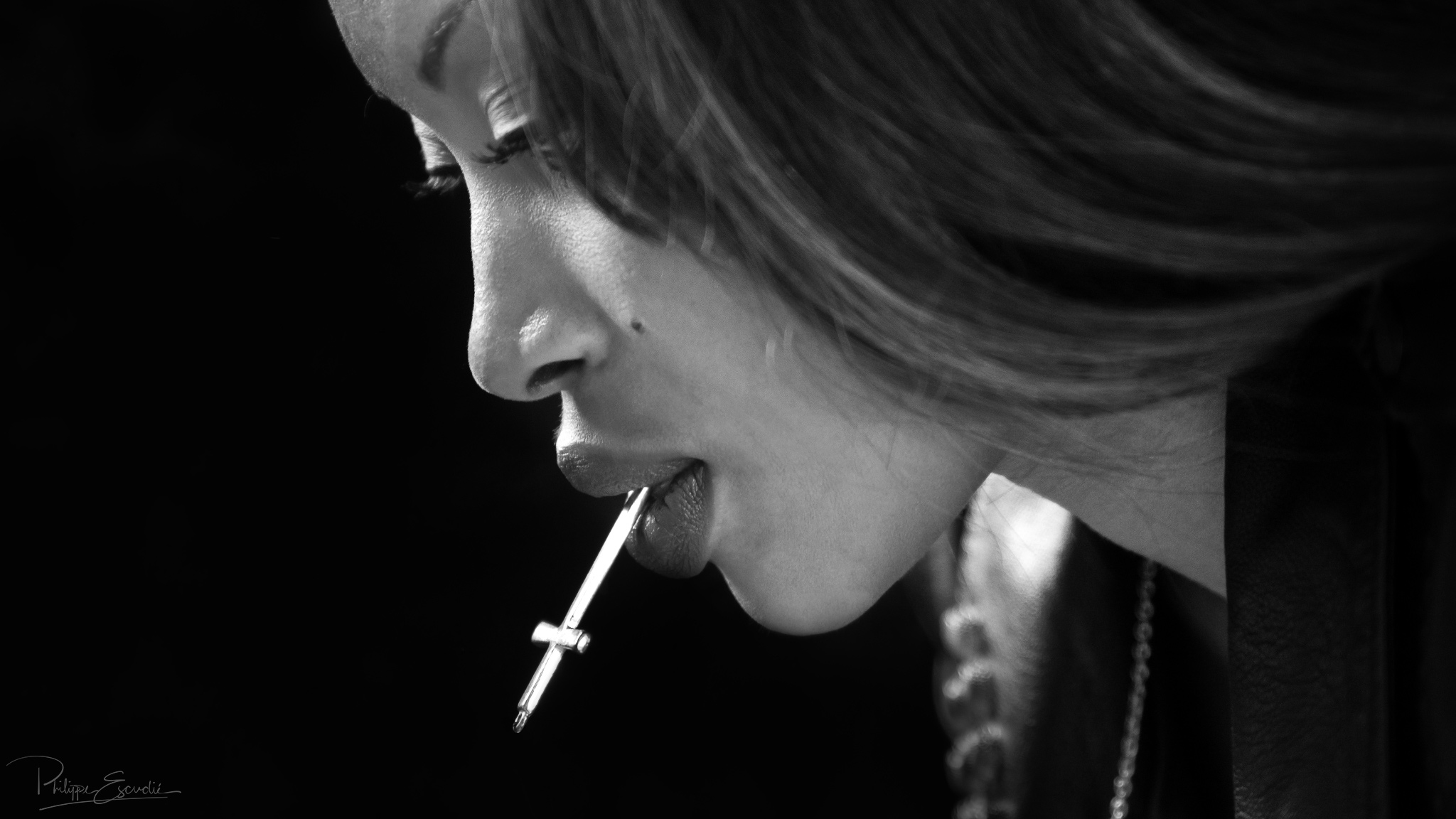 Gros plan du visage du modèle photo Nina, en noir et blanc, tenant une croix d'argent dans sa bouche
