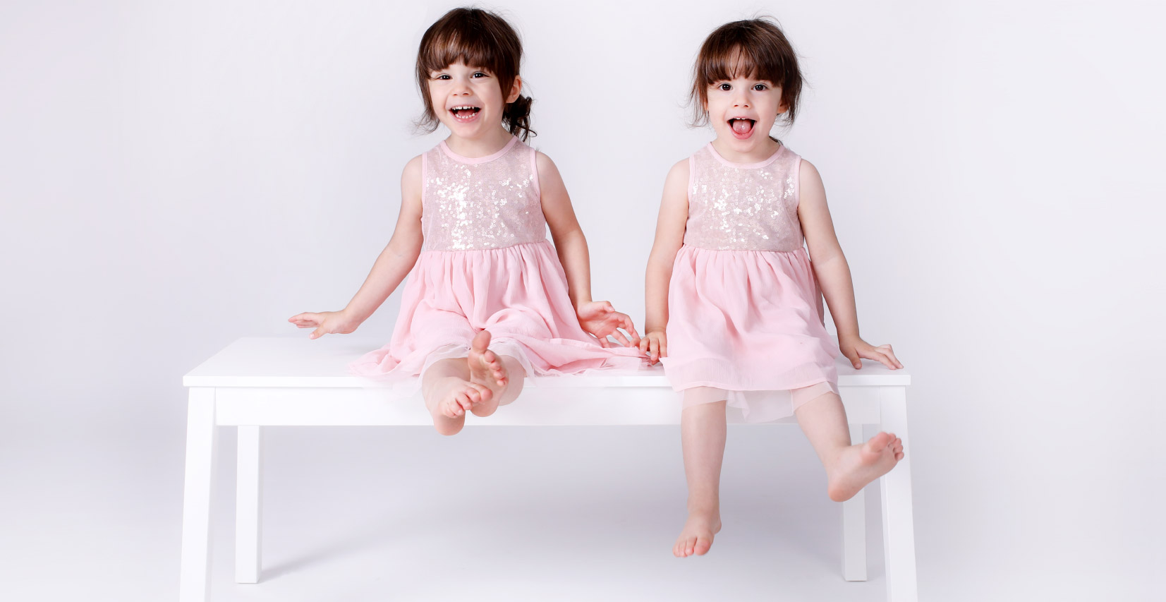 Deux petites filles jumelles souriantes en robe rose, assises sur un banc blanc dans un studio photo