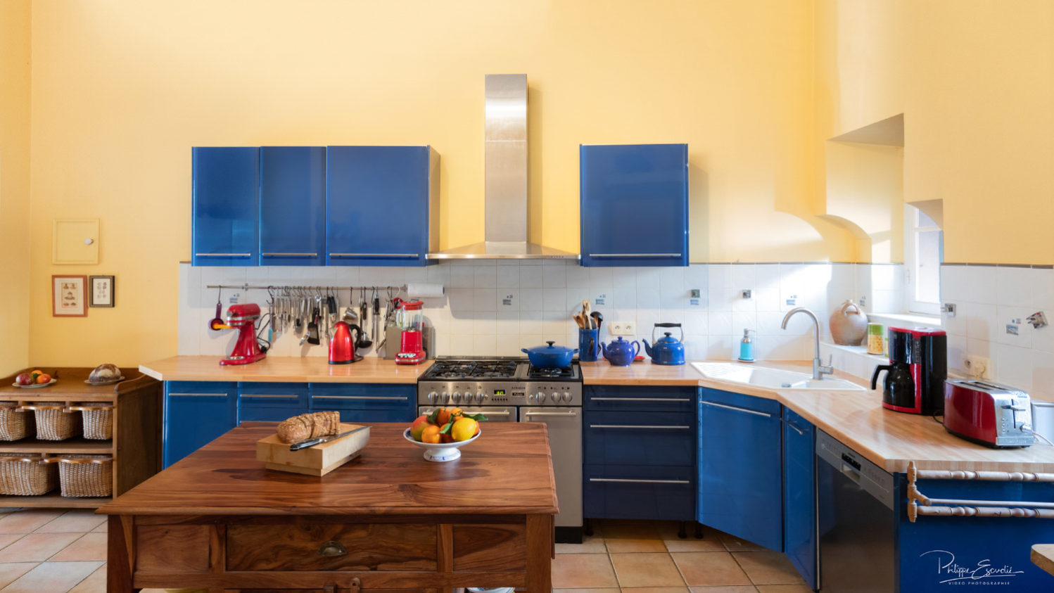 Cuisine de maison de maître, avec des placards bleus et une table en bois au premier plan sur laquelle reposent du pain et une corbeille de fruits.