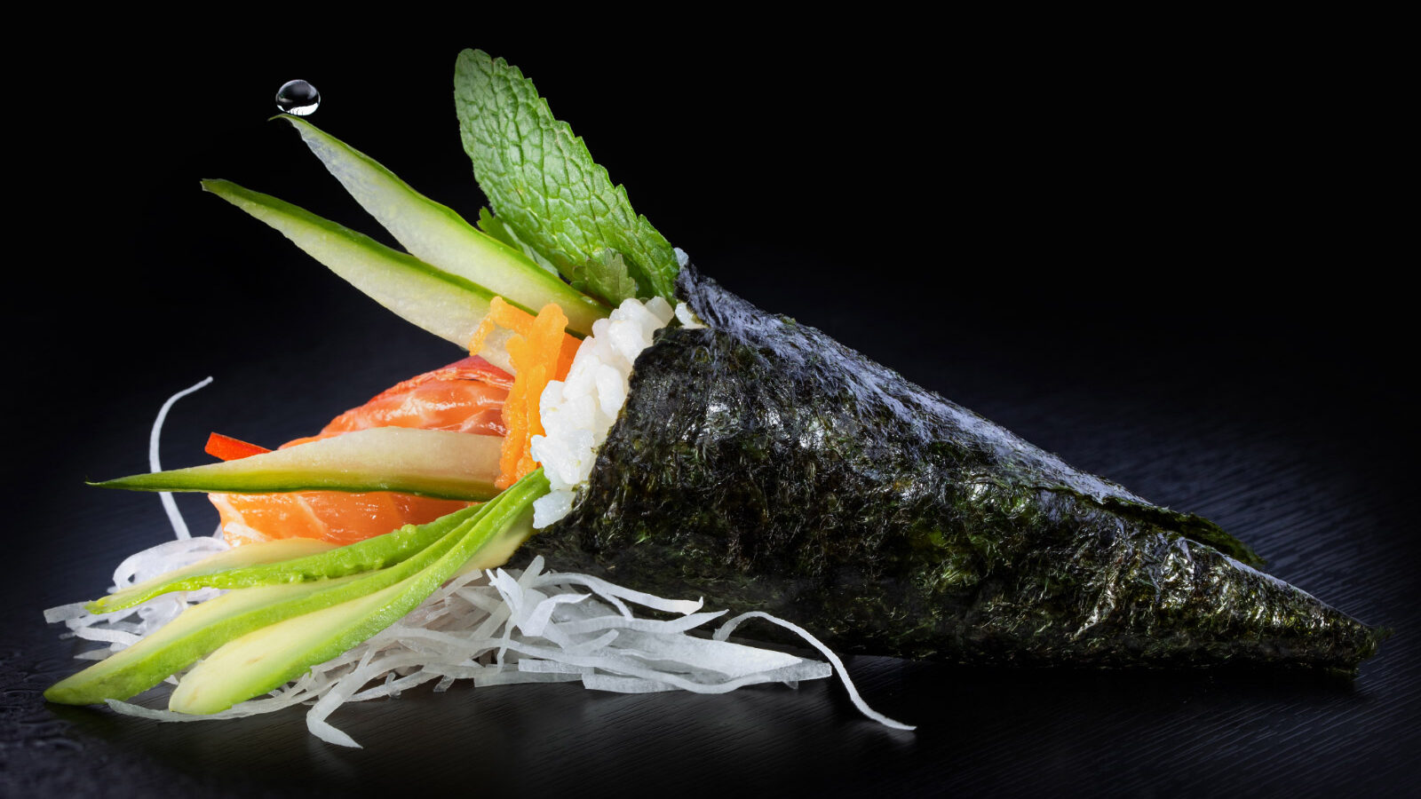 Un temaki saumon vu de profil sur une table noire avec une goutte d'eau qui tombe dessus.