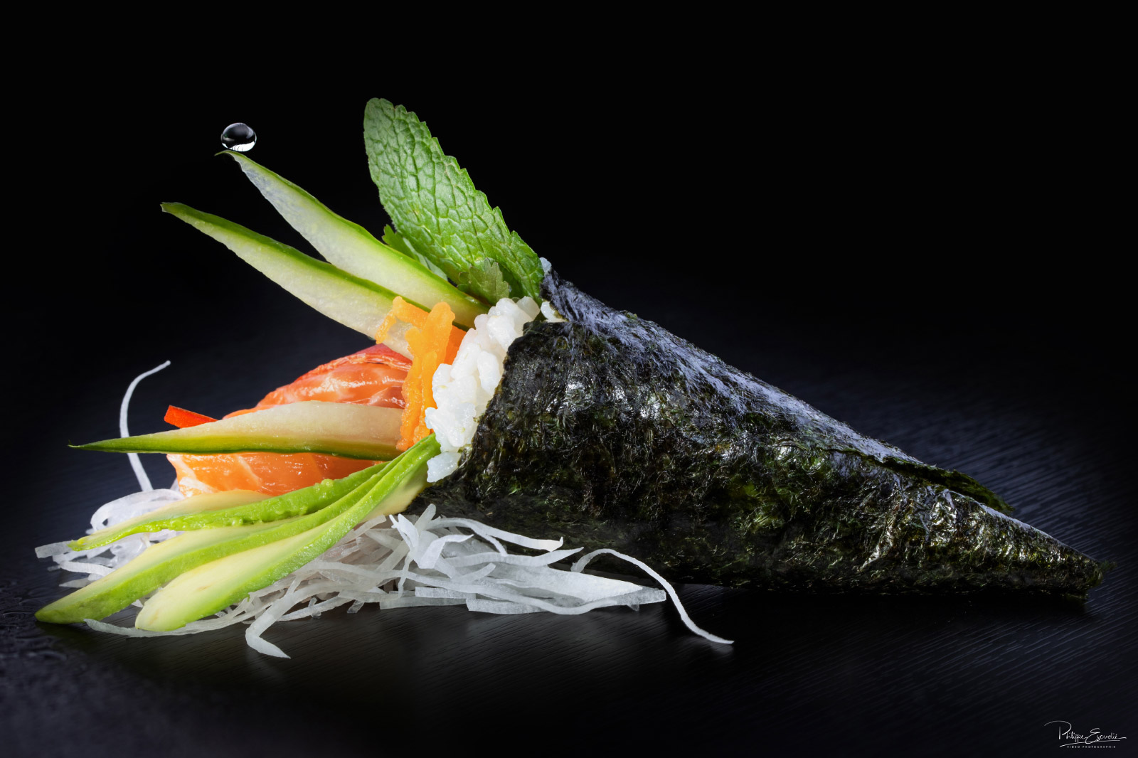 Un temaki saumon vu de profil sur une table noire avec une goutte d'eau qui tombe dessus.
